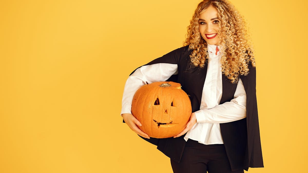 Smiling young woman in a Dracula costume holding a pumpkin against a vibrant yellow background.